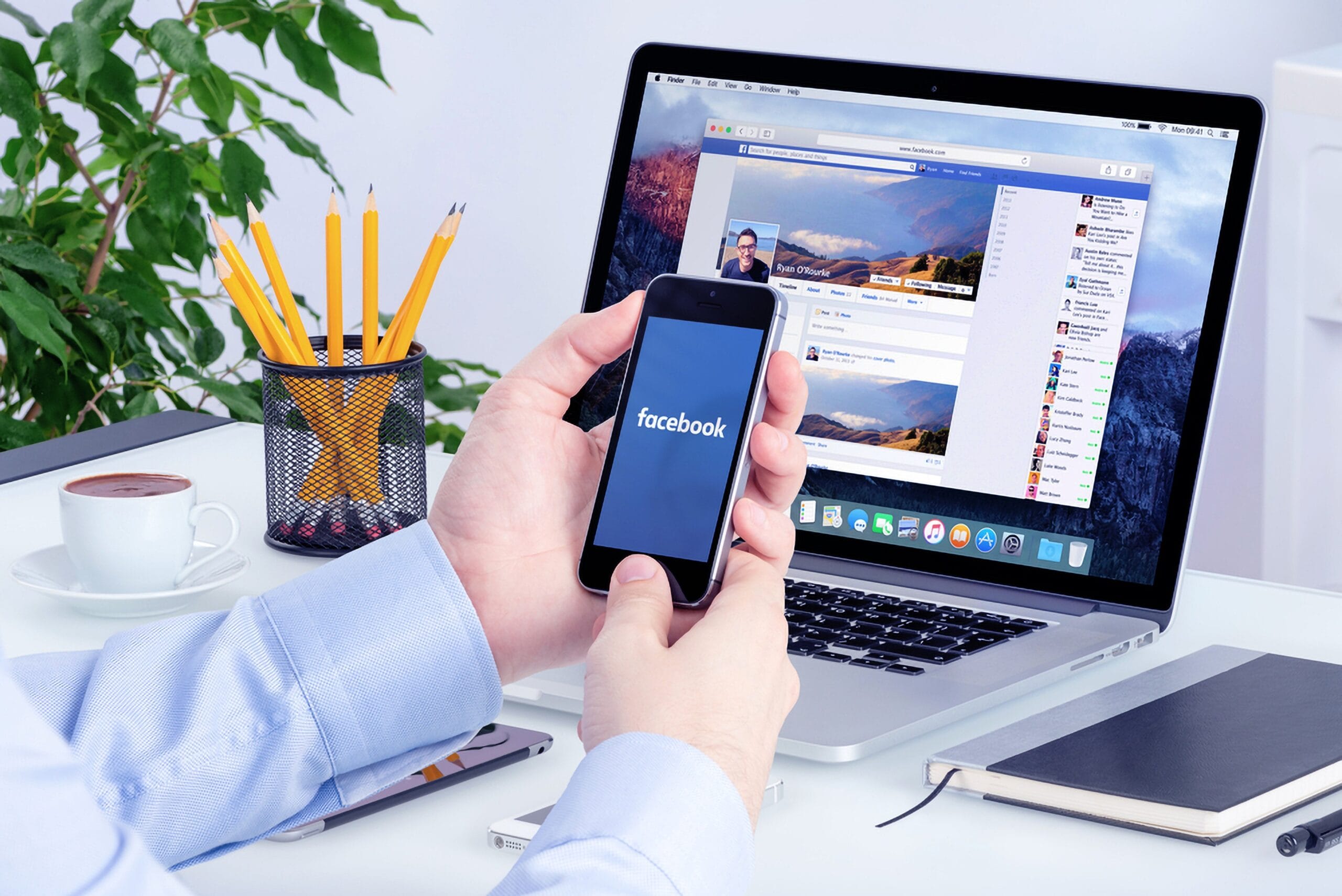 Virtual admin assistance services — Hand of a man in front of a laptop on a desk, holding his mobile phone with the Facebook app on the mobile screen. This depicts how important digital advertising like Facebook ads and Google ads are.